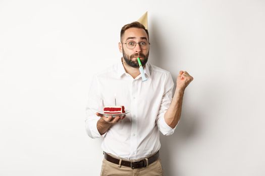 Holidays and celebration. Cheerful man enjoying birthday, blowing party whistle and holding bday cake, white background