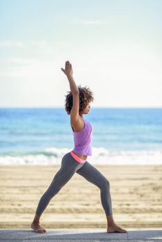 Black woman, afro hairstyle, doing yoga in warrior asana in the beach. Young Female wearing sport clothes with sea at the background
