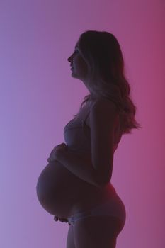 Beautiful young pregnant woman in lingerie is happily stroking her belly in studio in beautiful neon purple lighting. Concept of beauty and expectation of meeting with your beloved baby