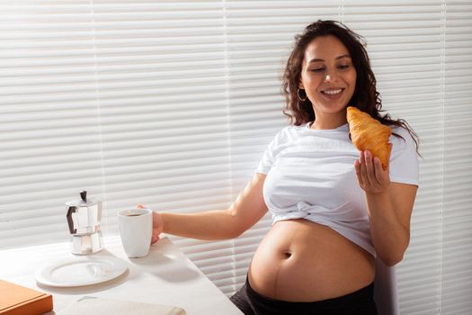 Joyful pregnant young beautiful woman eating croissant and drinking tea during morning breakfast. Concept of pleasant morning and expectation of meeting baby. Copyspace