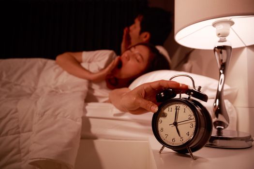 sleepy couple in bed with extending hand to alarm clock in the morning