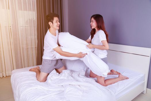 cheerful young couple having a bolster pillow fight on bed in bedroom