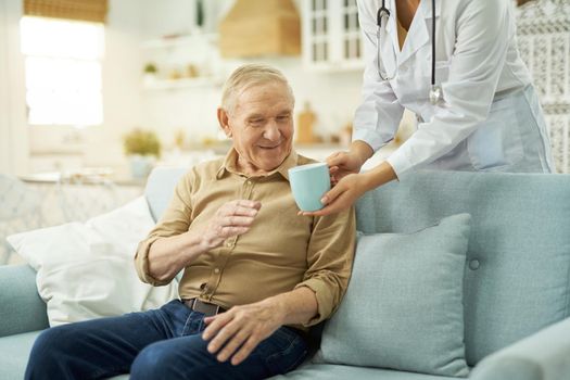 Caring nurse in white coat serving a cup to older man at his house while taking care of him