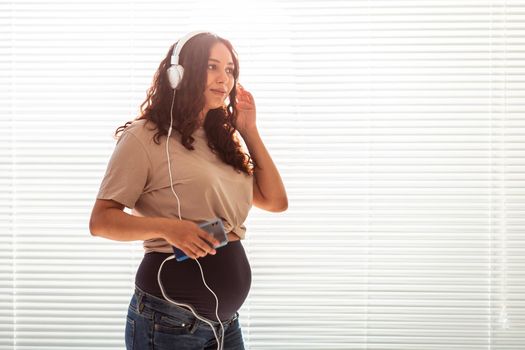 Beautiful young peaceful pregnant woman listens to pleasant classical music using smartphone and headphones. Concept of positive attitude before childbirth