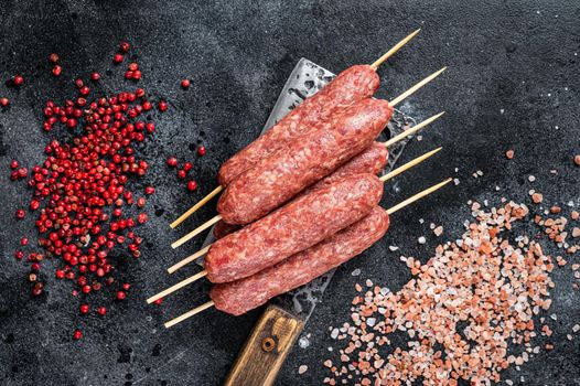 Raw lula or kofta kebabs skewers on a butcher cleaver with salt and pepper. Black background. Top view