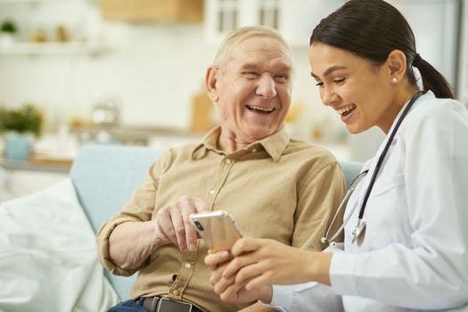 Happy nurse and senior citizen sitting on sofa and using mobile phone