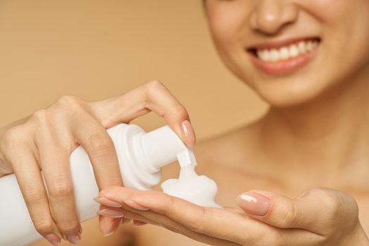 Cropped shot of young woman smiling, holding a bottle of gentle foam facial cleanser isolated over beige background