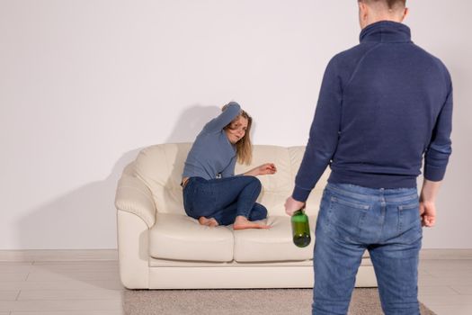 People, violence, alcoholism and abuse concept - Man with bottle alcohol while wife is lying on sofa