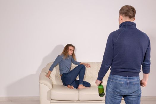 People, violence, alcoholism and abuse concept - Man with bottle alcohol while wife is lying on sofa
