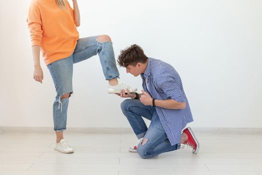 Young man is kneeling and reverently tying shoelaces to his domineering unidentified woman posing on a white background. Concept of dominant relationships.