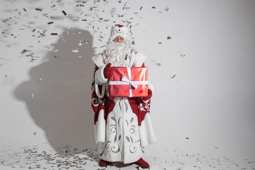 father frost in long warm coat, red mittens and a hat holds a gift with a lot of confetti around him