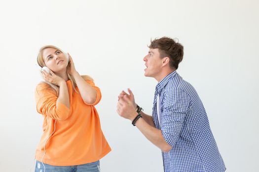 Young upset man begs his woman to listen to him but she listens to music with headphones posing on a white background. Misunderstanding and unwillingness to engage in dialogue.