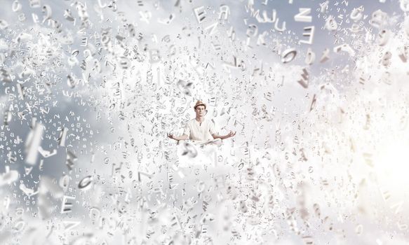 Man in white clothing keeping eyes closed and looking concentrated while meditating among flying letters in the air with cloudy skyscape on background.