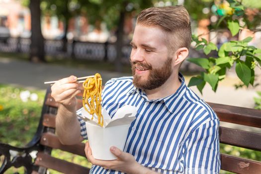 Side view of a young handsome hipster guy eating chinese noodles from a lunch box while sitting in a park bench on a sunny summer day. The concept of a healthy and nourishing snack.