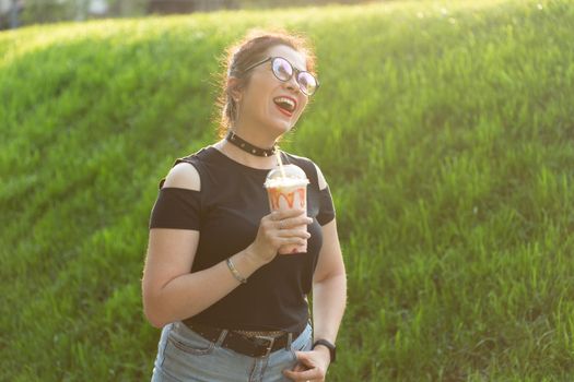 Portrait of a positive young pretty girl in punk clothes and glasses with a milkshake in her hands posing in a summer park on a warm summer evening. Vacation getaway concept.