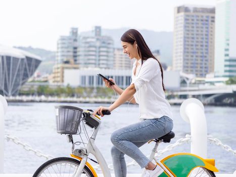 Smiling girl in headphones, holding smartphone and riding bicycle in the park