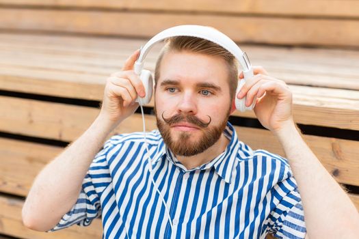 Portrait of a handsome young student guy with a mustache and a beard relaxing in a park with headphones on a warm summer day. Weekend getaway concept.
