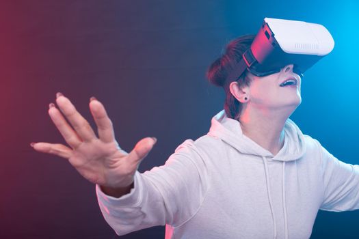Passionate middle-aged woman in sweater is watching new film with glasses of virtual reality on red-blue background. Concept of high technology accessibility for middle-aged people. Advertising space.