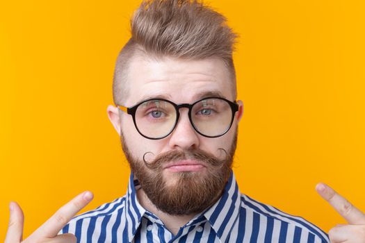 Charming confident young fashion hipster man with glasses and a beard shows on himself posing over a yellow background. Place for advertising. The concept of self-confidence and success.