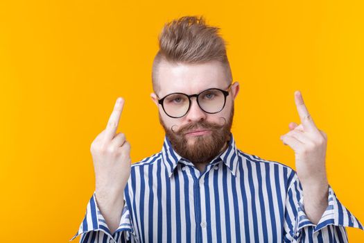 An angry young stylish man in glasses with a mustache and beard shows middle finger posing on a yellow background. Concept of denial and annoyance.