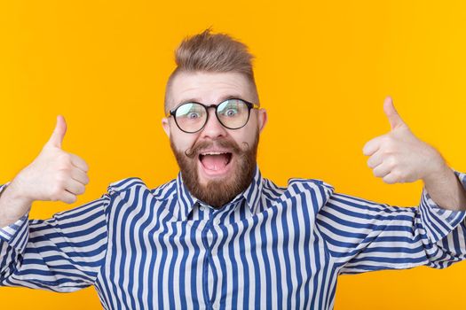 Joyful handsome young hipster man with a mustache and beard posing on a yellow background showing thumbs up. Telephony and communications concept.