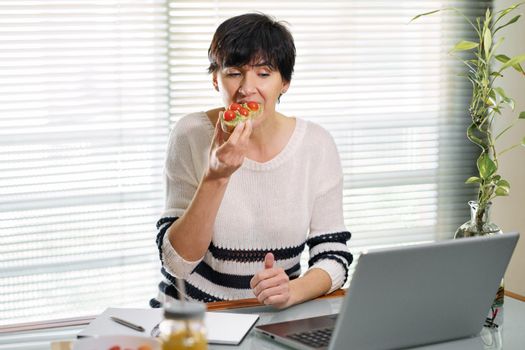 Woman eating some healthy food, while teleworking from home on her laptop.