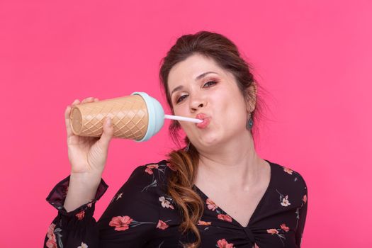 Portrait of a beautiful brown-eyed young woman drinking cocktail with a straw while posing on a bright pink background. Concept of summer and desserts.