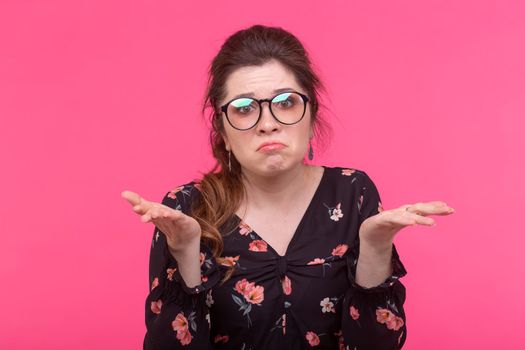 Close-up portrait of a sad disappointed young woman with glasses waving her arms posing on a pink background. Concept of disappointment and loss.