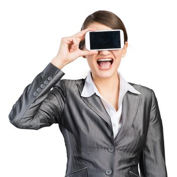 Portrait of happy woman covering her eyes with smartphone. Businesswoman showing mobile phone with blank screen. Corporate businessperson isolated on white background. Mobile communication layout