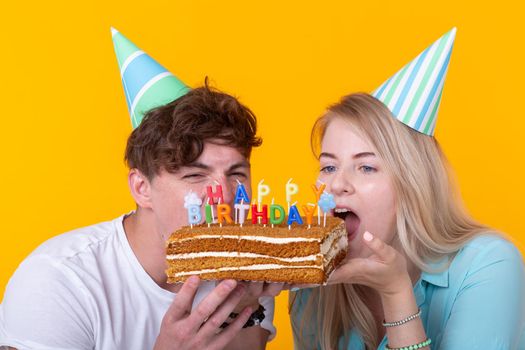 Close up of bizarre young couple girl and boy in paper hats want to bite off a piece of congratulatory cake. Birthday and congratulations concept.