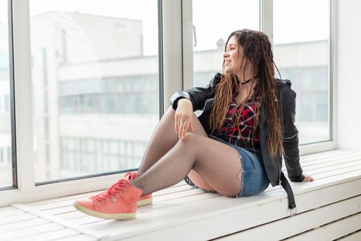 Young stylish woman with dreadlocks sitting on the windowsill