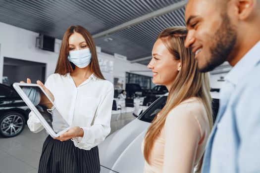 Car saleswoman wearing medical mask shows buyers couple something on digital tablet