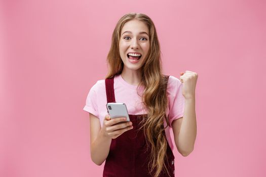 Yes I did it. Happy young excited cute woman in trendy outfit expressing cheer and joy smiling happily at camera raising fist in triumph and success holding smartphone beating own record in game