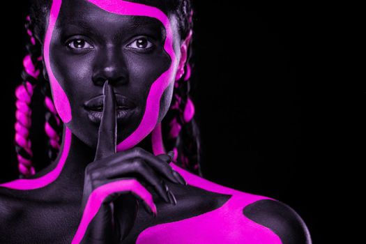 Face art. Girl showing gesture quietly, asks remain silent with finger. Woman with black and pink body paint. Young african girl with colorful bodypaint. An amazing afro american model with makeup.