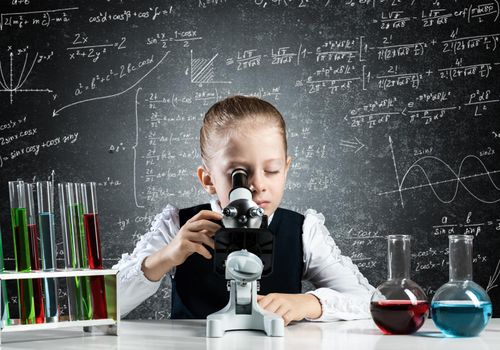 Little girl scientist looking through microscope in classroom at chemistry lesson. School chemical laboratory with glass flasks and test tubes. Schoolgirl making experiment in chemistry class.