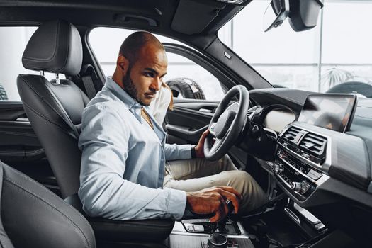 Young african american man sitting in a new car in car showroom and looking around