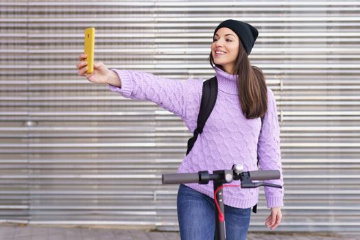 Woman in her twenties with electric scooter taking a selfie with a smartphone outdoors.