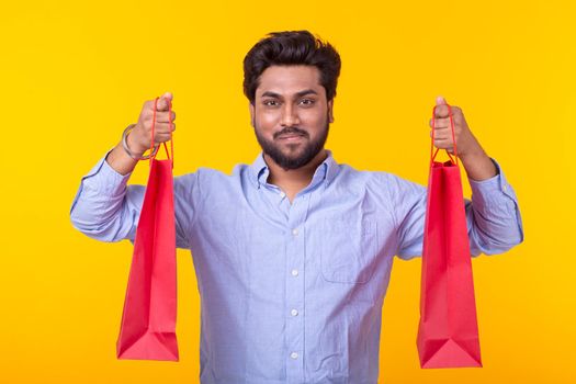 Young handsome indian man with a beard is holding red shopping bags posing on a yellow background. Supermarket shopping concept.