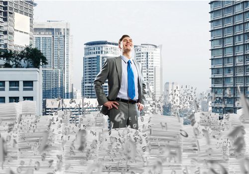 Confident young businessman in suit standing among flying letters with cityscape on background. Mixed media.
