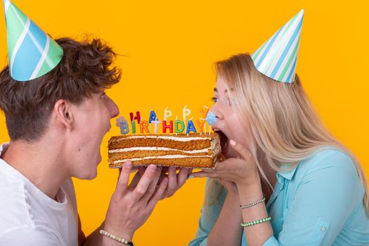 Close up of bizarre young couple girl and boy in paper hats want to bite off a piece of congratulatory cake. Birthday and congratulations concept.