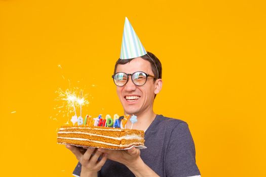 Crazy cheerful young man in glasses and paper congratulatory hats holding cakes happy birthday standing on a yellow background. Jubilee congratulations concept.