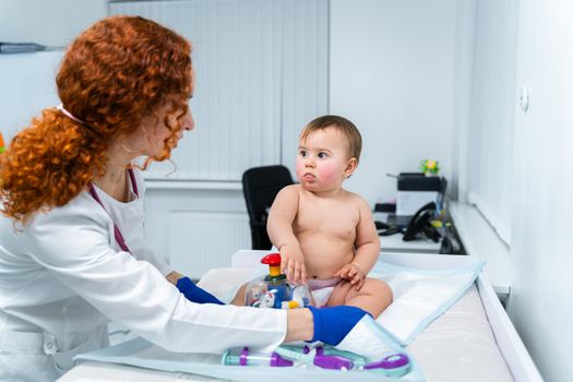 Red-haired female doctor examines little child for less than a year in a modern clinic. Pediatrician during examination baby in the hospital. Childrens healthcare. Diagnostics and treatment for kid