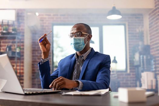 Business man with face mask thinking about marketing strategy and using laptop in office