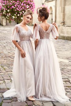 Two beautiful bridesmaids ladies in gorgeous elegant stylish light grey silver floor length open back dresses in old beautiful European city on a wedding day.