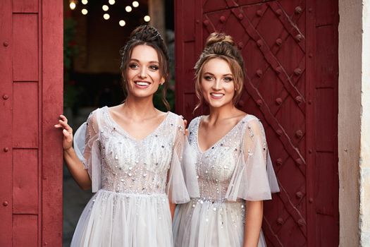 Beautiful retro styled bridesmaids ladies in gorgeous elegant stylish light grey silver floor length dresses in old beautiful European city welcome greeting guests at the gate.