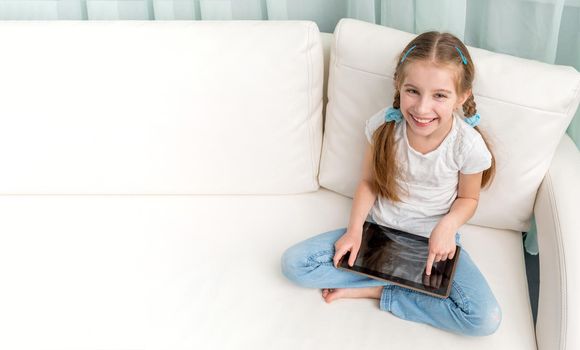 cheerful little girl with tablet on her legs looking at camera