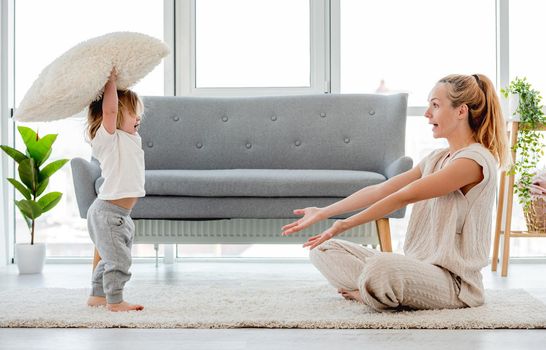 Beautiful blond young mother sitting on the floor and playing with her baby child son with pillow. Happy family portrait