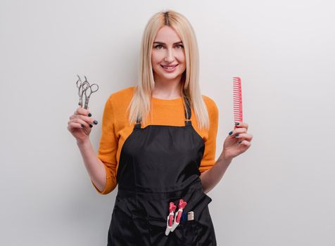 Blond woman hairdresser smiling and holding in her hands professional scissors and comb isolated on white background. Closeup portrait