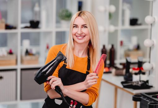 Beautiful blond girl hairdresser looking at camera, smiling and holding hair dryer at beauty salon