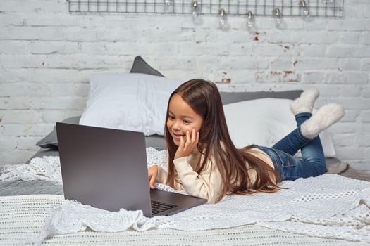 Cute little girl girl feeling amusing while watching cartoons on a laptop sitting on bed